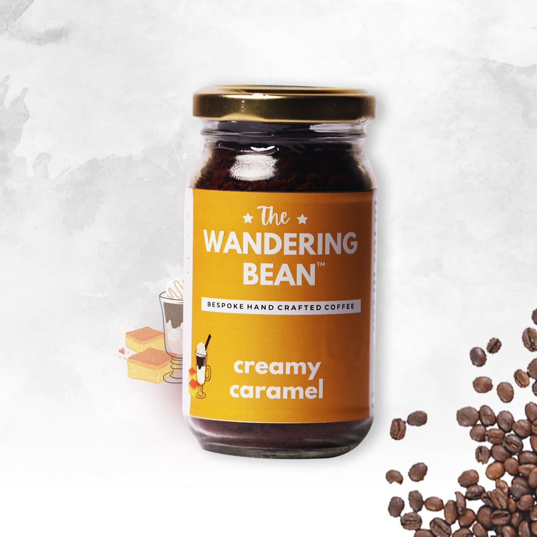 A jar of The Wandering Bean instant coffee with the label 'Creamy Caramel' prominently displayed. The jar is placed in front of a backdrop featuring coffee beans, with a small illustration of a cup and a piece of chocolate to the left.