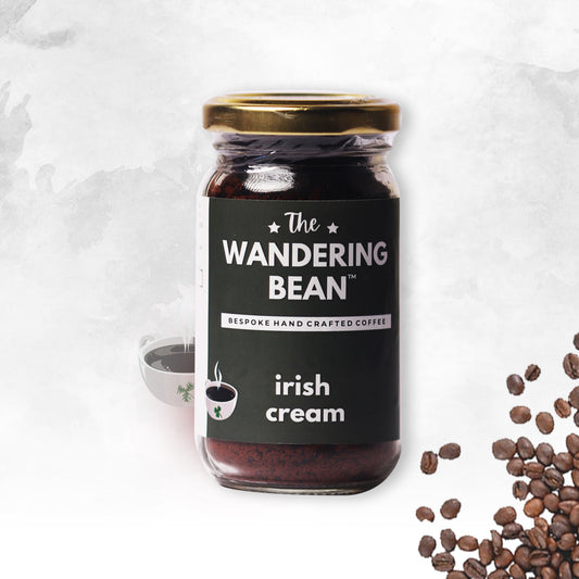 A jar of 'The Wandering Bean' Irish Cream Instant Coffee, with a label that states 'REPLAY HAND-CRAFTED COFFEE'. There is a cup of coffee in the foreground, and coffee beans scattered around the background.